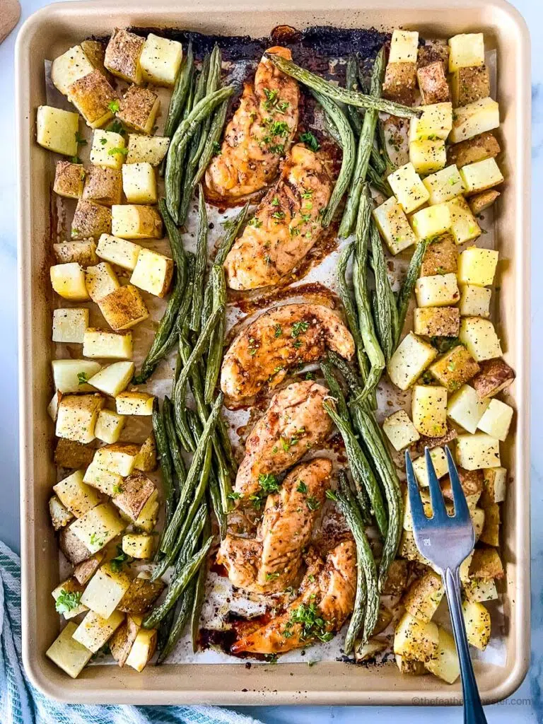 a baking sheet with oven baked marinated chicken tenders, asparagus, and potatoes with a fork at the side.