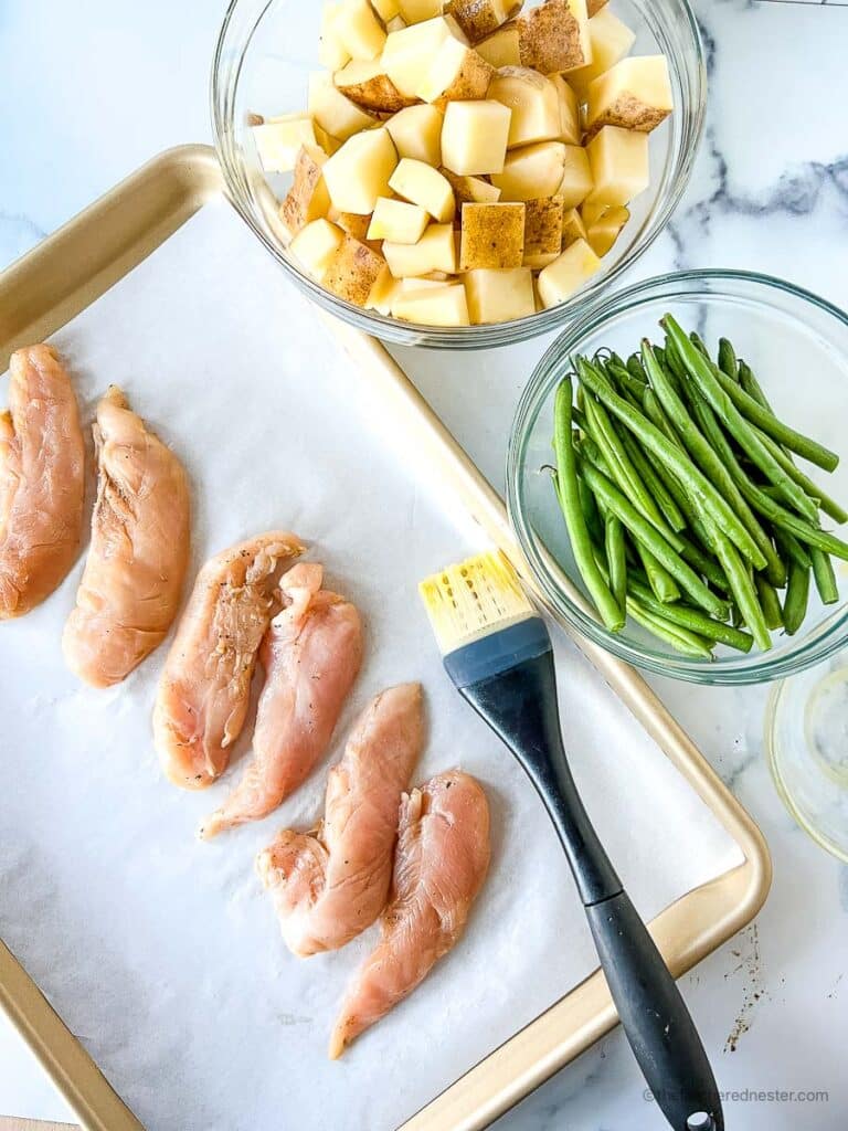 chicken tenders lined in a parchment paper placed in a baking sheet with a basting brush in it, and a bowl of potatoes and asparagus on the side. 