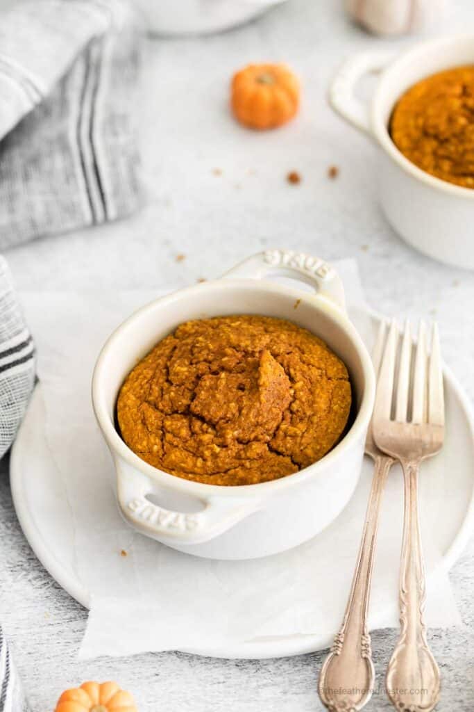 a  white ramekin with pumpkin baked oats with forks on the side and place on top of a white plate with a gray cloth and another ramekin with oats in the background.