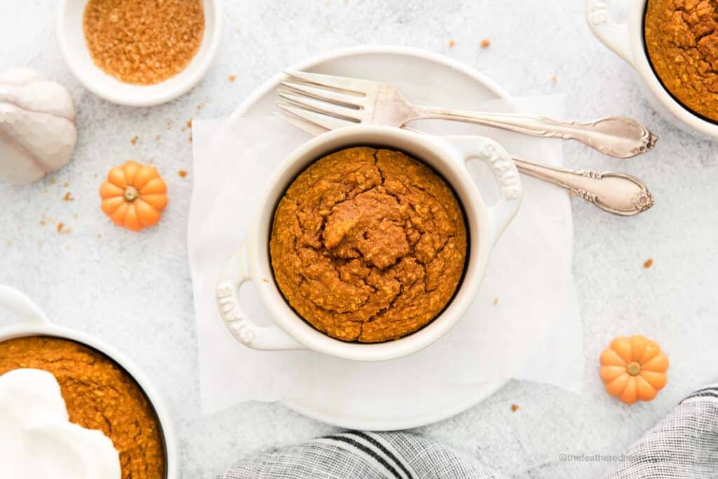 landscape view photo of a white ramekin with pumpkin baked oats with forks on the side and place on top of a white plate with a gray cloth and another ramekin with oats in the background.