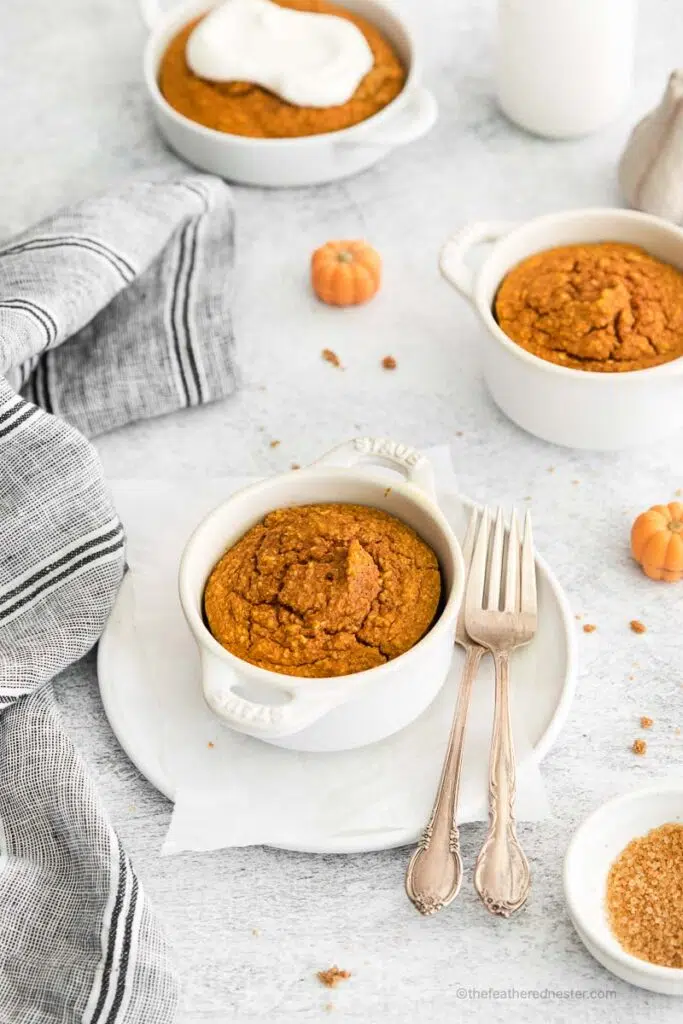 a white ramekin with pumpkin baked oats with forks on the side and place on top of a white plate with a gray cloth and another bowls of ramekin with oats in the background.