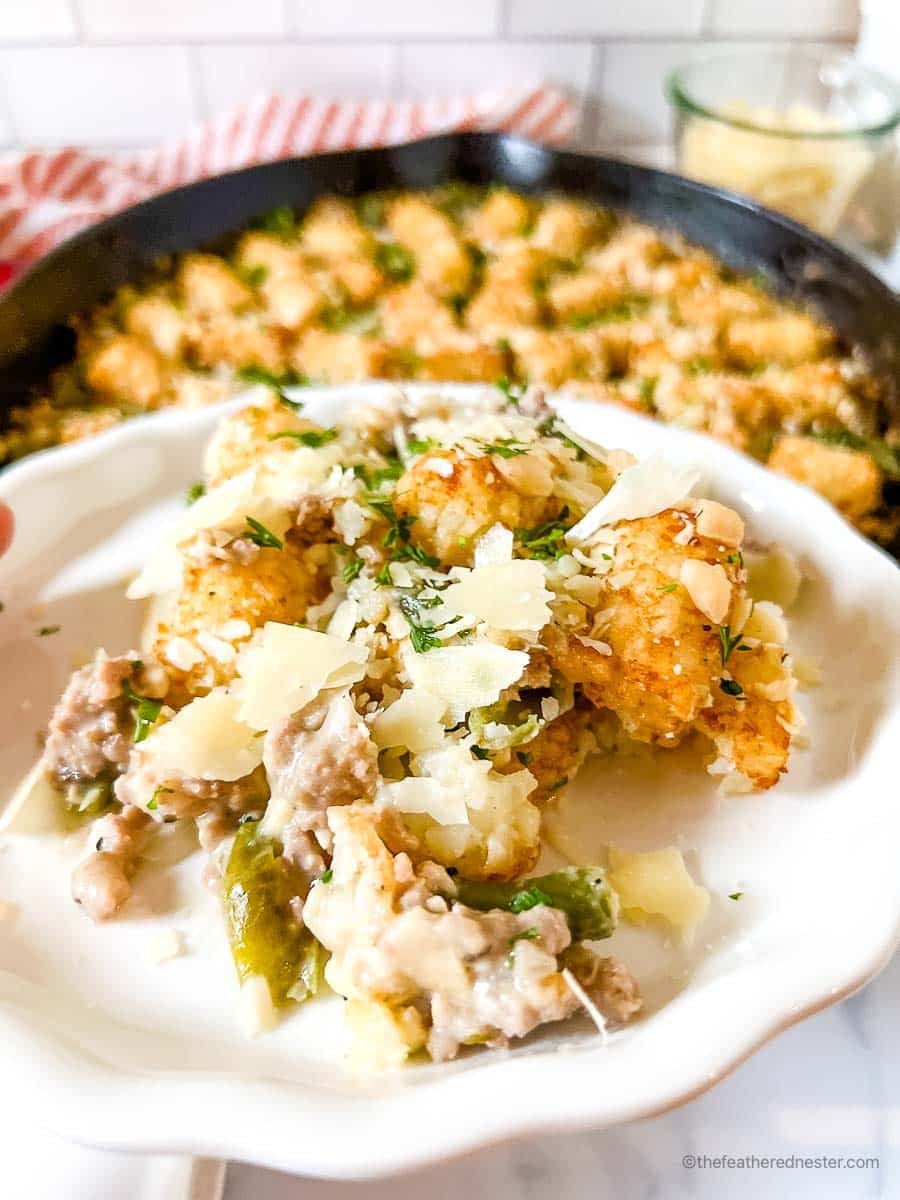 a white plate with a serving of turkey tater tots with skillet, red striped cloth, and a jar of shredded cheese in the background.