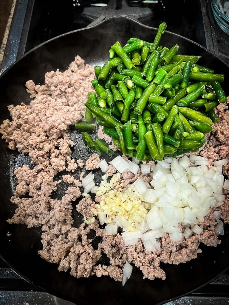 green beans, onions, and garlic are added to the browned ground turkey in a cast iron skillet. 