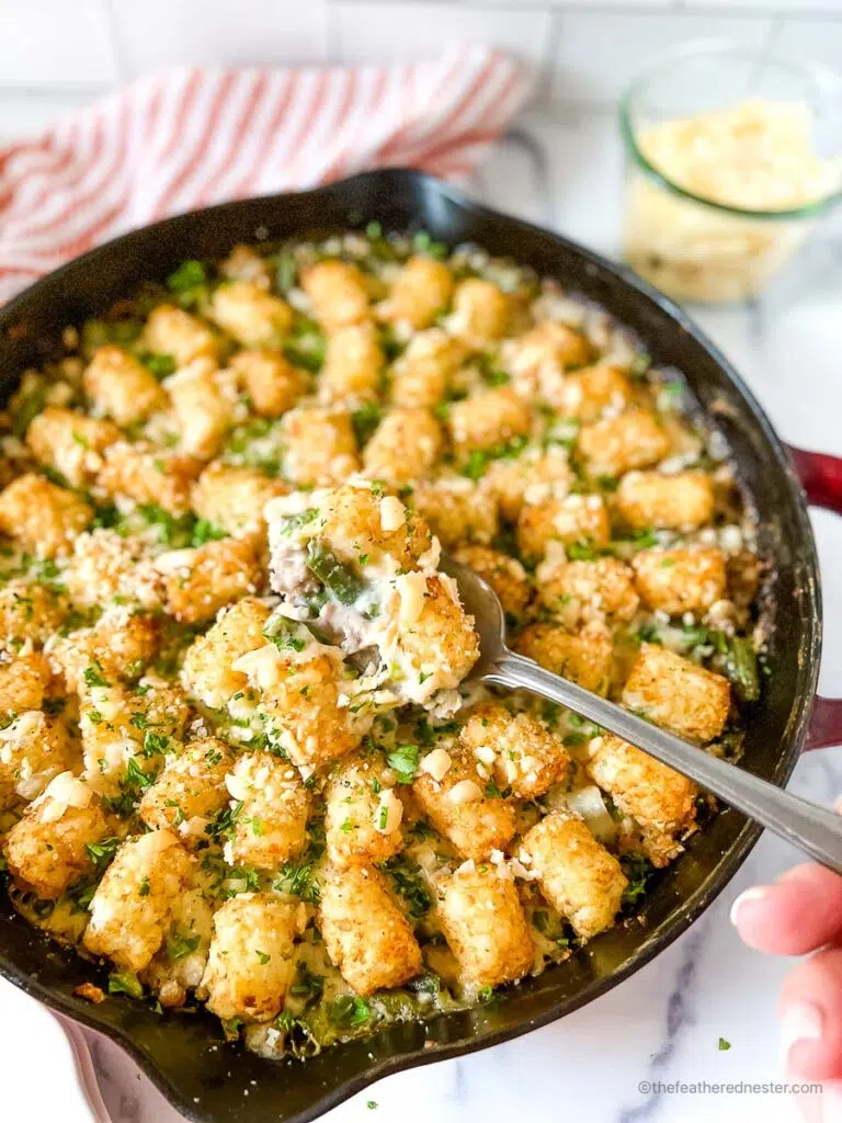 Turkey Tater Tot Casserole in a cast iron skillet with a hand holding a spoon scooping the turkey tater tots and a red striped cloth, and a jar of shredded cheese in the background.