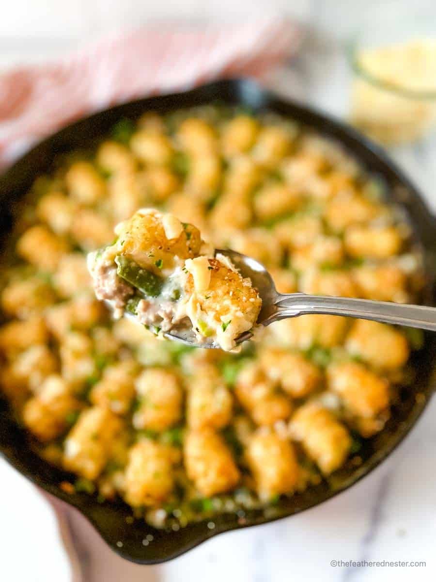 a spoon with a scoop of turkey tater tot casserole and a skillet full of tater tots, striped red cloth, and a jar of shredded cheese in the background.