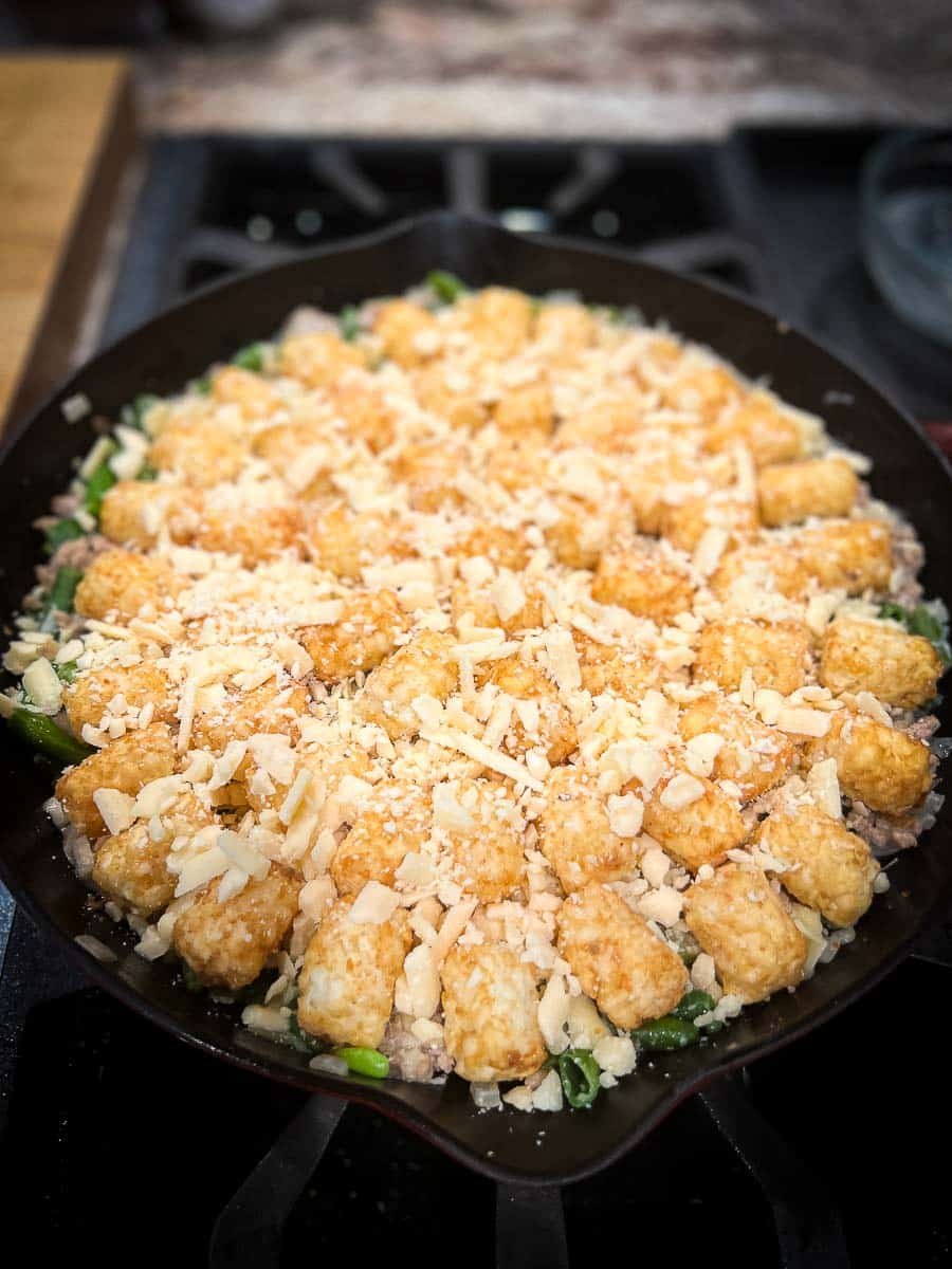 tater tots and cheese layered on top of the ground beef mixture in a cast iron skillet.