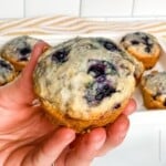square photo of a hand holding a Bisquick blueberry muffin with more muffins at the back.