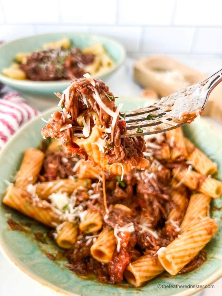 a fork with pasta coated with braised short rib ragu and two green bowls with short rib rigatoni, red striped cloth and a salt and pepper cellar at the back.