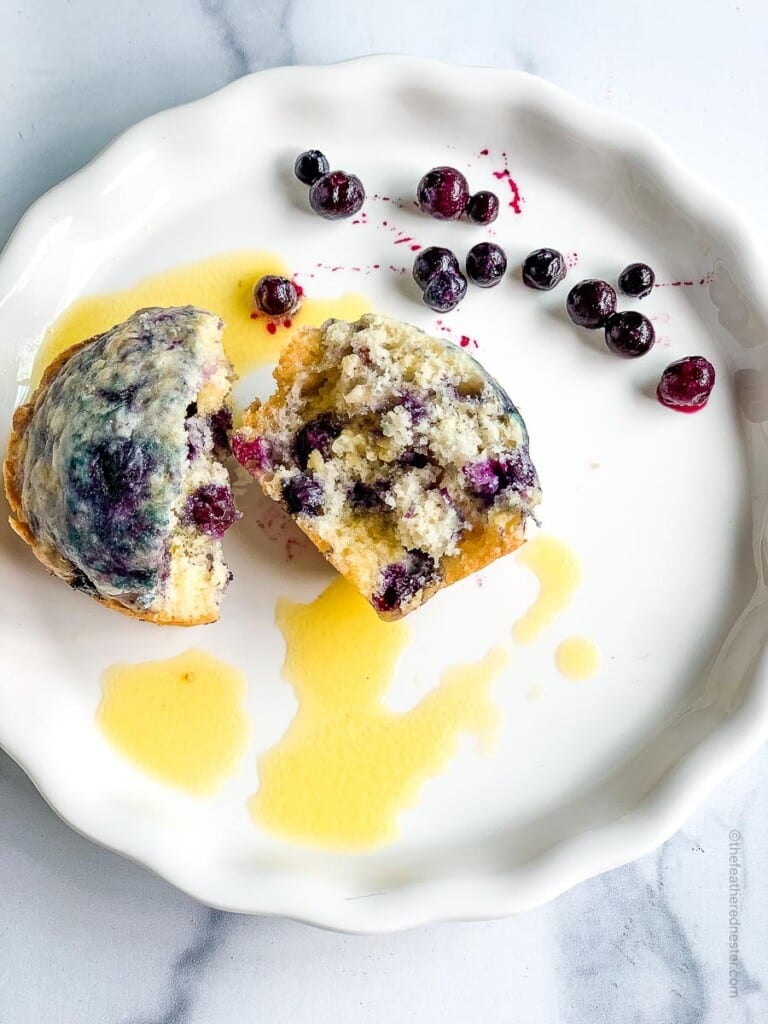 tender blueberry muffin cut in half, drizzled with melted butter, sitting on a plate next to some fresh berries.