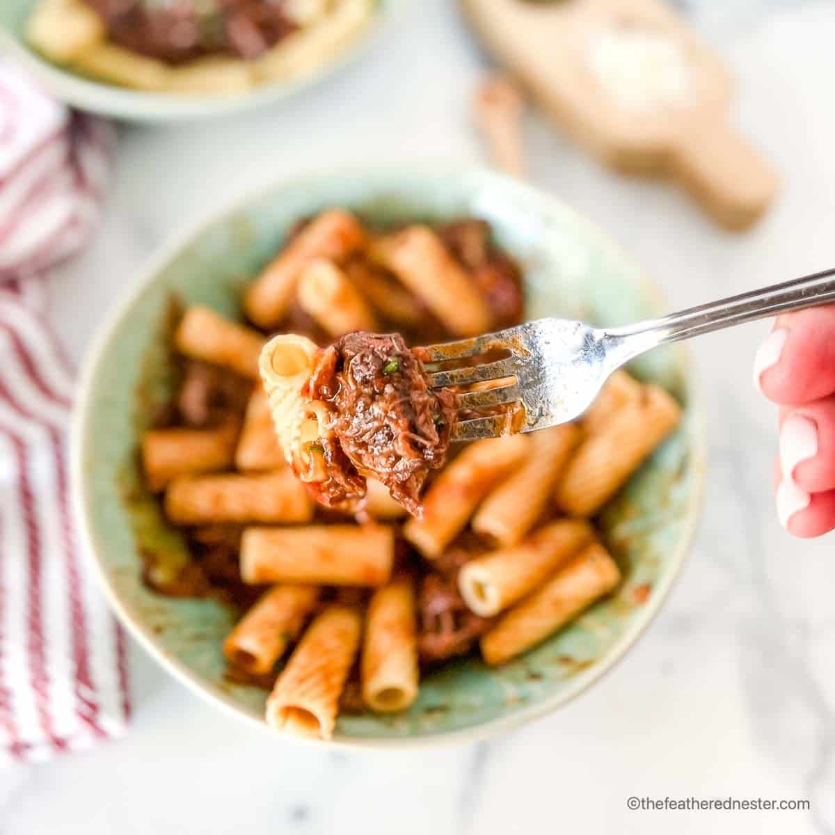 a fork with pasta coated with braised short rib ragu and a green bowl with short rib rigatoni at the back.