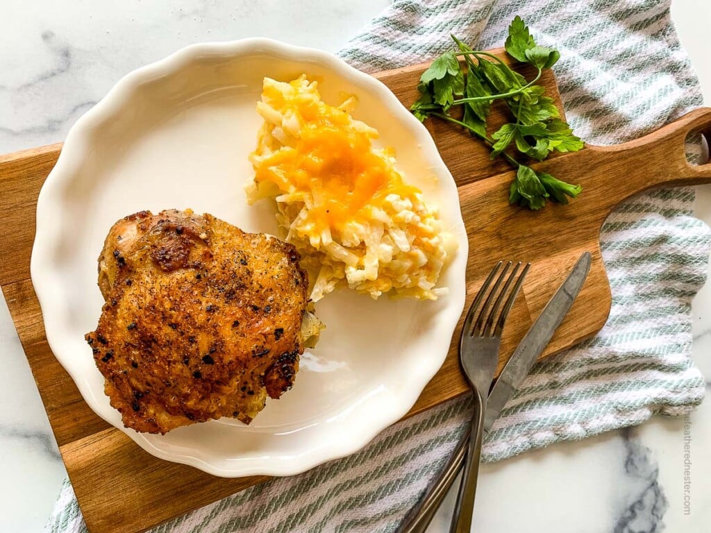 horizontal photo of a white plate with cast iron skillet chicken and hash brown placed on top of a wooden board.
