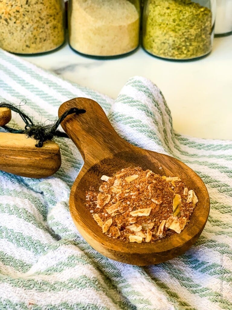 a wooden measuring spoon with chicken taco seasoning with striped kitchen cloth and seasonings in the background.