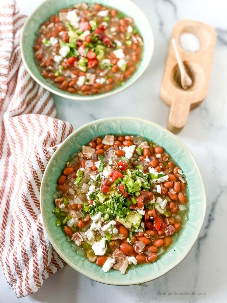 two green bowls of crock pot pinto beans with ham. A striped red cloth and salt and pepper cellar sit on the side.