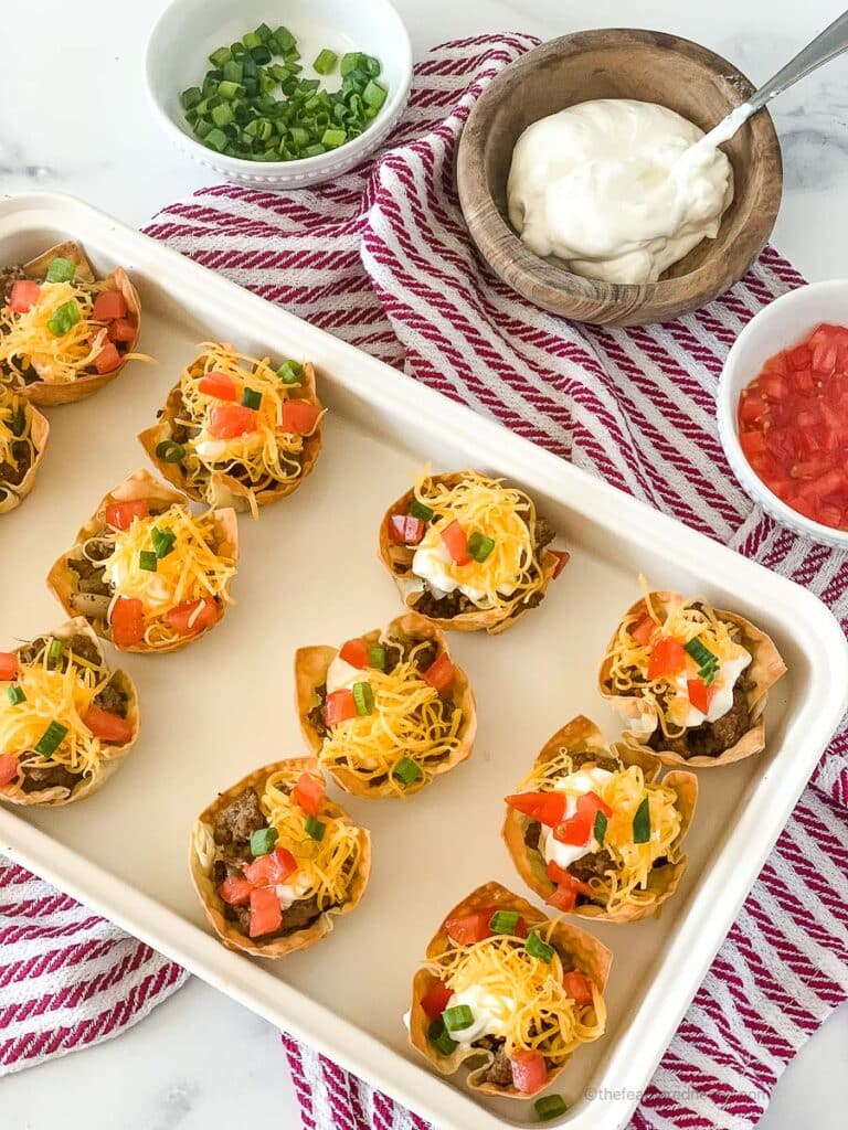a white tray of taco wonton cups on top of a red and white napkin with bowls of cilantro, sour cream, and salsa