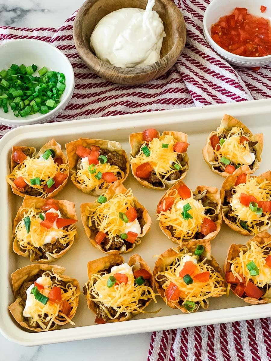 a white tray of taco wonton cups on top of a red and white napkin with bowls of cilantro, sour cream, and salsa