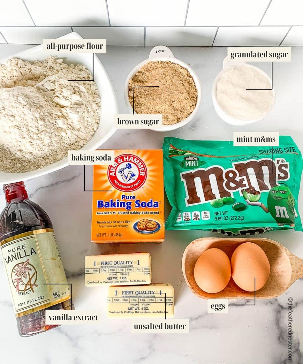 Ingredients on a counter top to make M&M christmas cookies:  mint-flavored candies, eggs, butter baking soda, brown sugar, flour, sugar, vanilla