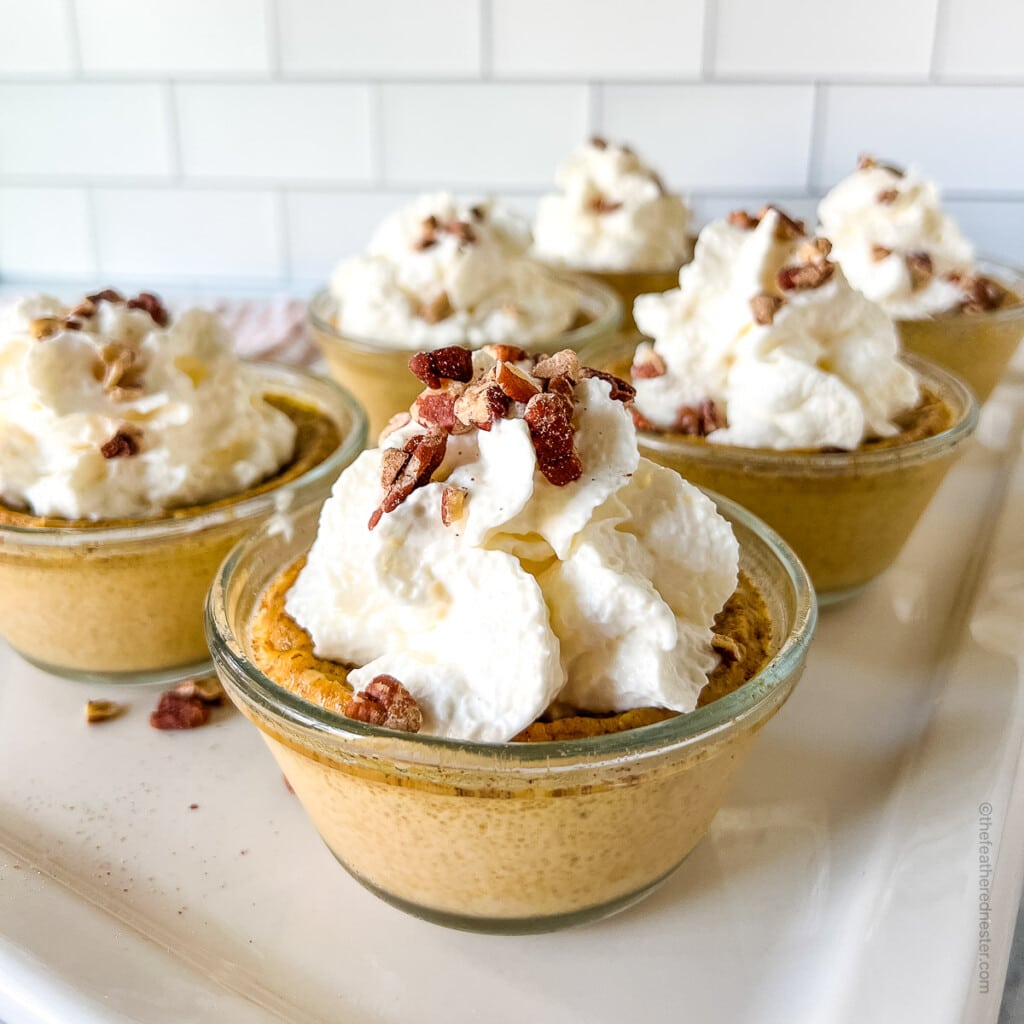 square photo of six cups of pumpkin custard and with whipped cream and nuts on top.
