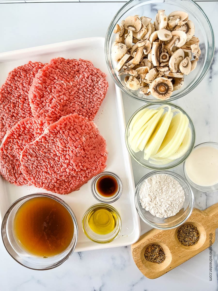 Ingredients in bowls for a cube steak recipe, including beef shoulder steaks, Montreal steak seasoning, cooking oil, yellow onions, white mushrooms, all purpose flour, beef broth, Worcestershire sauce, and heavy cream.