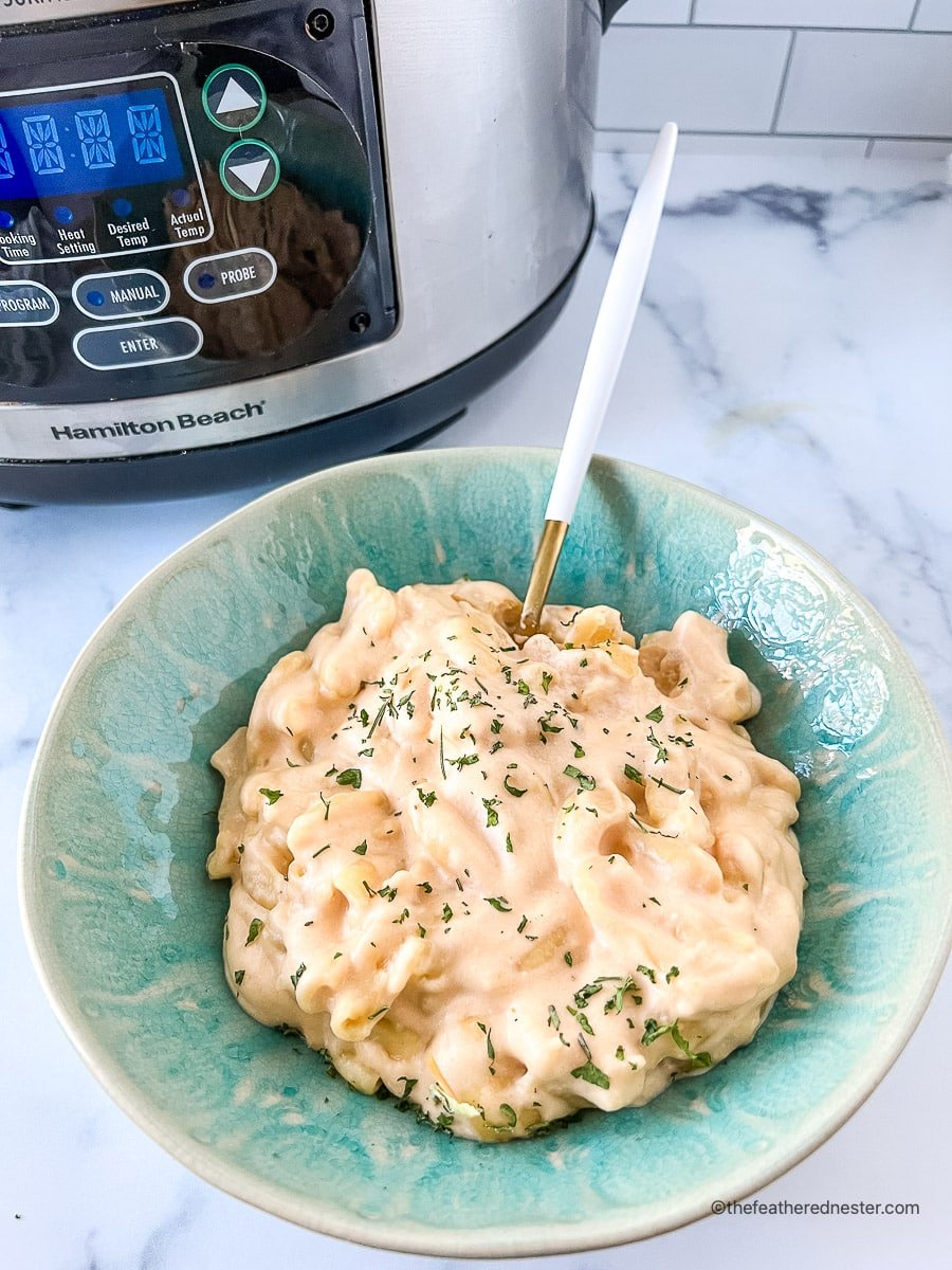 Spoon in a green bowl of 3 cheese mac and cheese with a Hamilton Beach crock pot in the background.