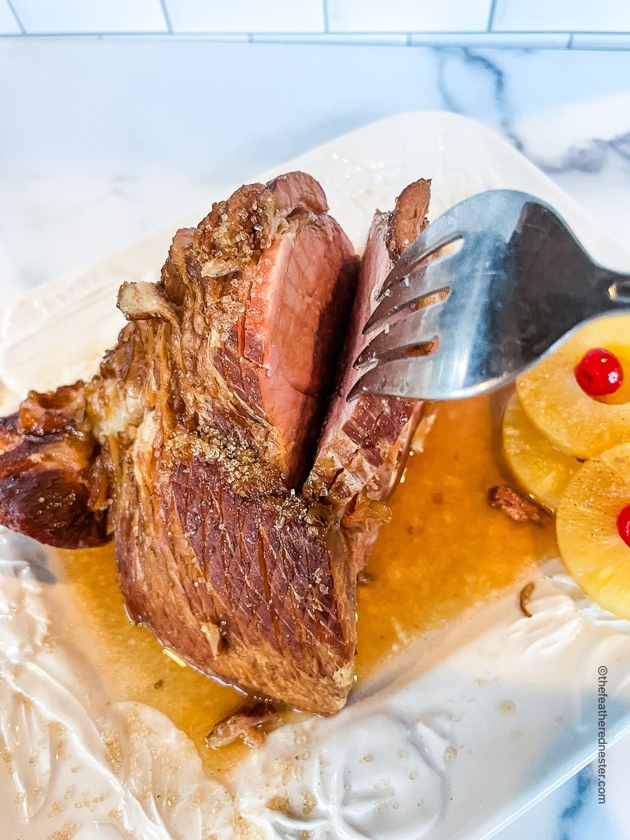 glazed and cooked Dr Pepper glazed ham on a plate with pineapple slices and a carving fork.