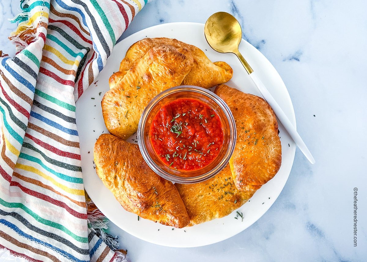 A plate of mini pepperoni calzones with dipping sauce in the middle and a gold spoon on the side.