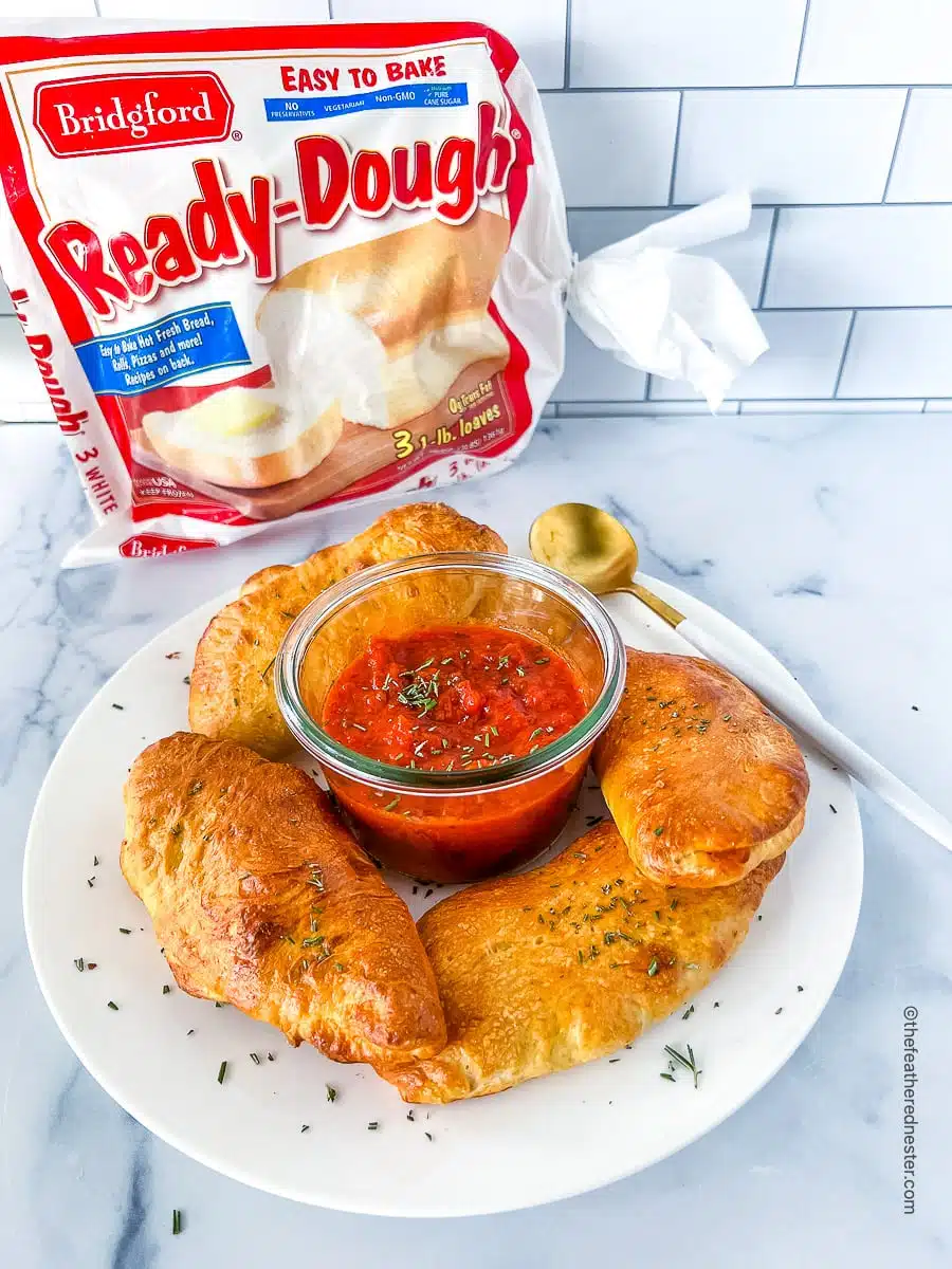 a white plate with Mini Pepperoni Calzones and marinara dipping sauce at the center, and a bag of Bridgford's Ready-Dough at the back.