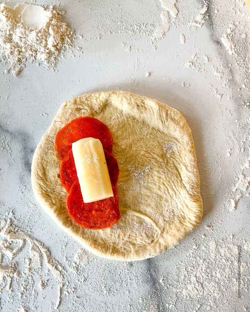 flattened dough with 3 pieces of pepperoni and a cut of string cheese on top of it.
