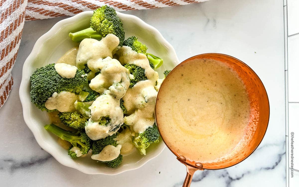 brocolli on a plate getting drizzled by white cheese sauce.