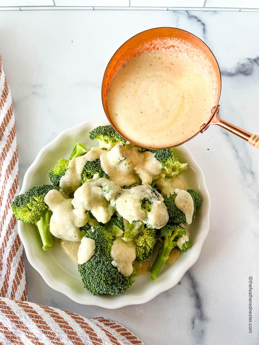 A bowl of broccoli being drizzled with a spoonful of white cheese sauce.