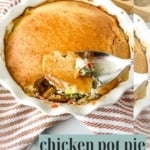 A spoon lifting a serving of chicken pot pie with a golden-brown crust from a white ruffled baking dish.