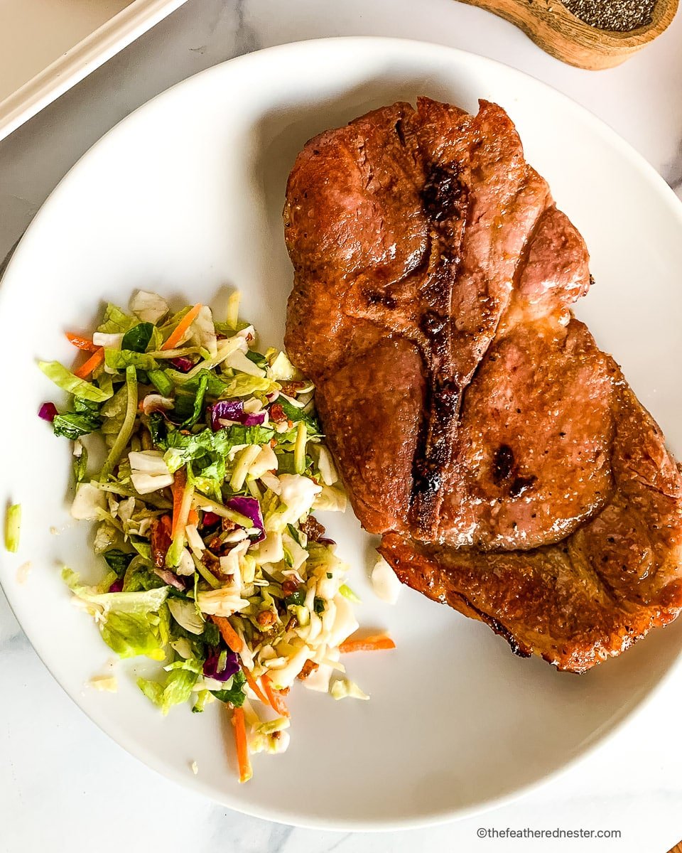 Juicy baked pork steak on a round dinner plate with serving of chopped vegetable salad.