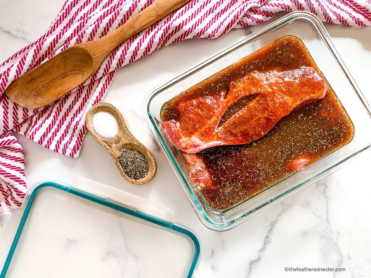 A glass pan with pork and marinade with a dish of salt and pepper 