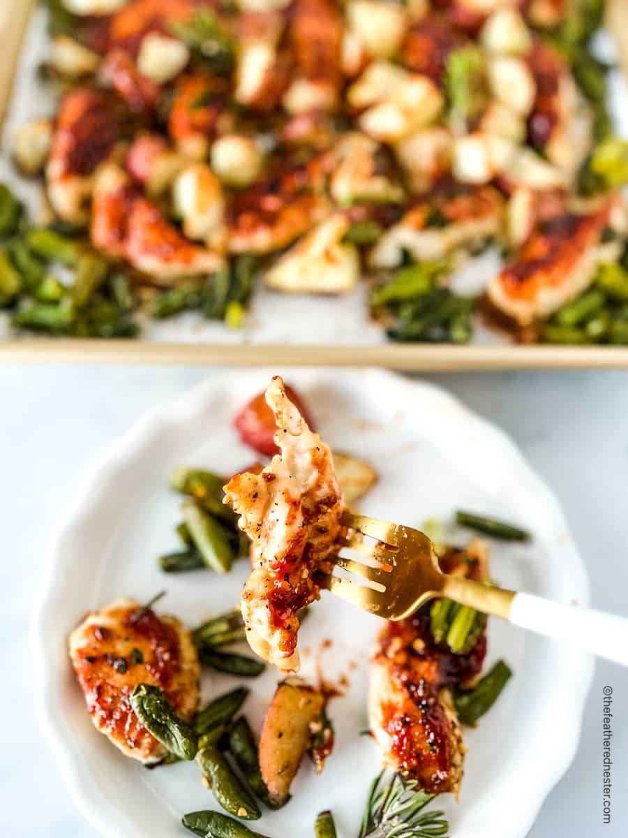 BBQ chicken tenders on a fork with remaining dinner on a plate under it, and sheet pan of oven baked BBQ chicken behind it.