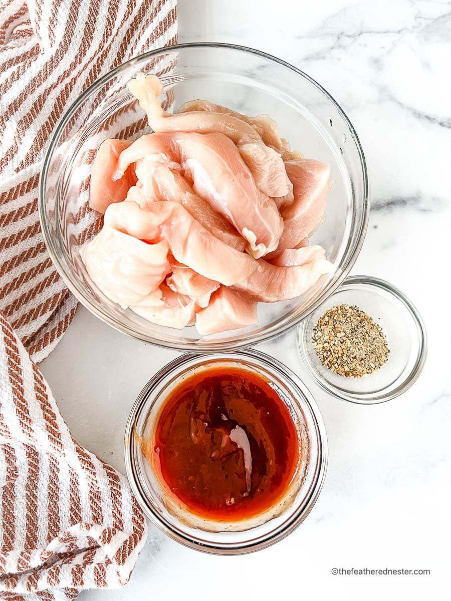 Raw chicken tenders in a glass bowl next to two small bowls with all-purpose seasoning and barbecue sauce.