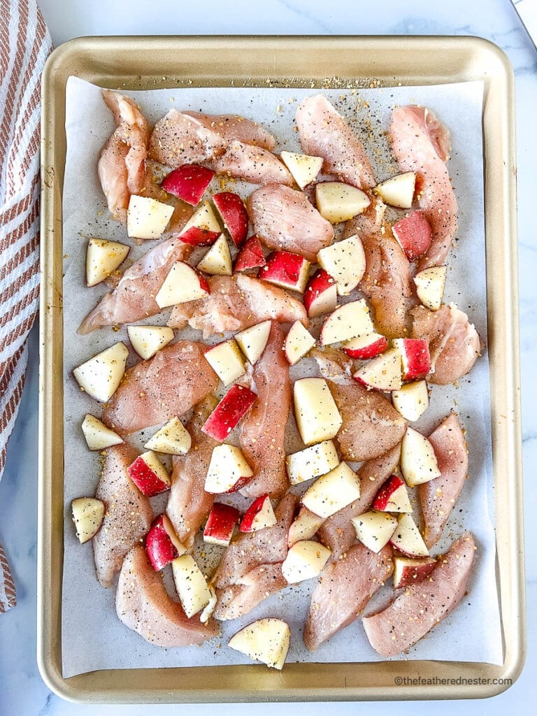 Slices of raw boneless skinless chicken breast and diced red potatoes on a sheet pan.