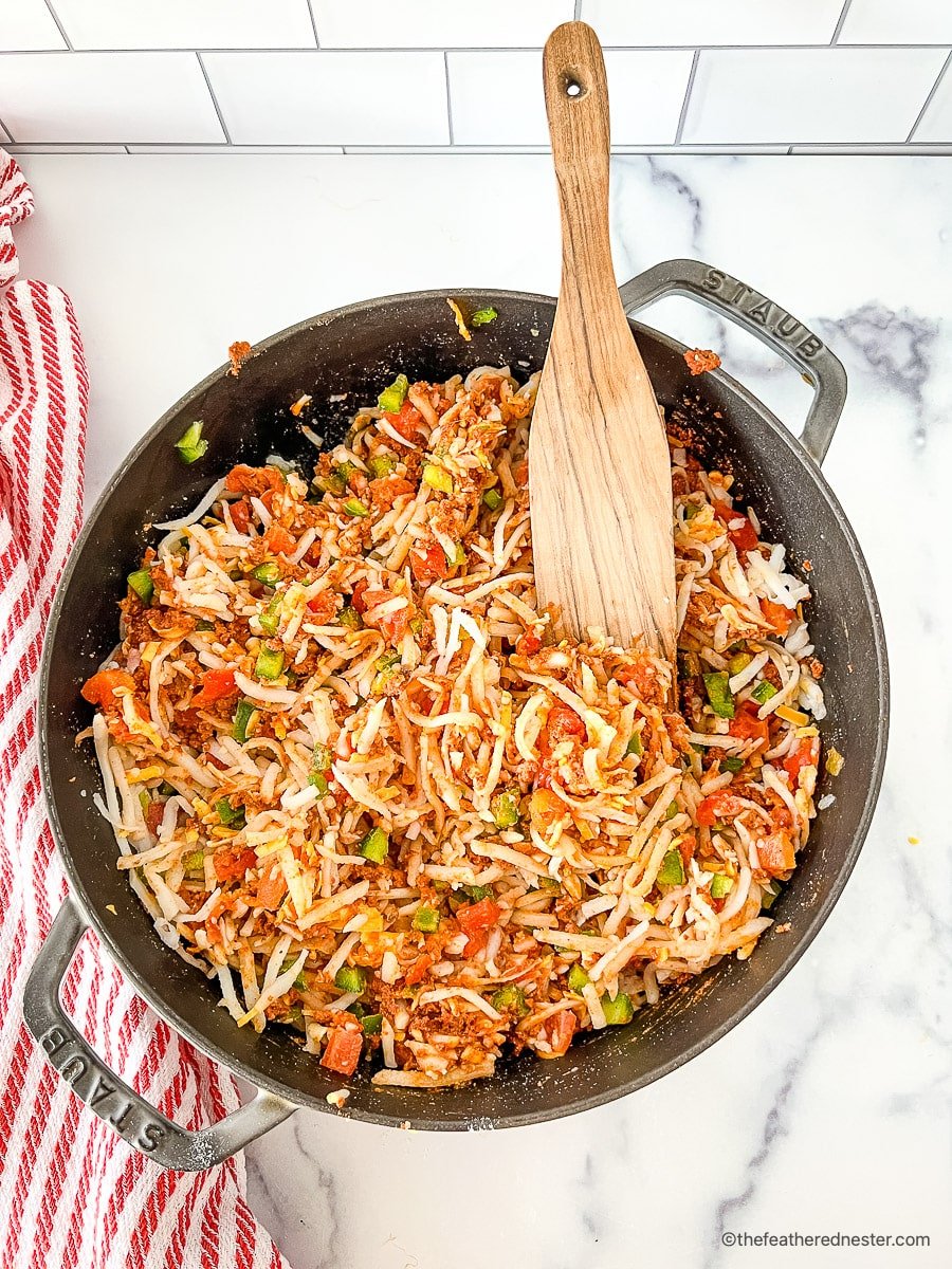 Wooden spoon in pot with shredded hash browns, diced vegetables and other ingredients for Tex Mex egg bake.