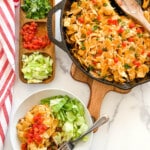 Frito casserole with dishes of shredded lettuce, tomatoes, and cilantro on a table. A plated serving in front.