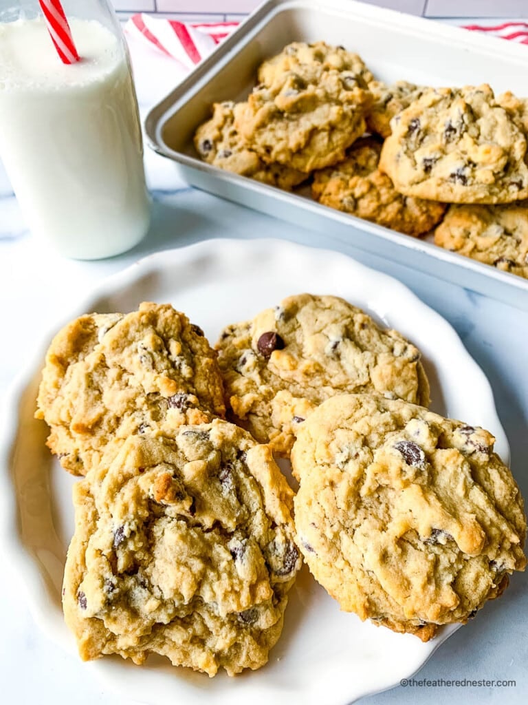 Four eggless chocolate chip cookies on a small white plate next to a glass of milk, with remaining batch of cookies on a platter.