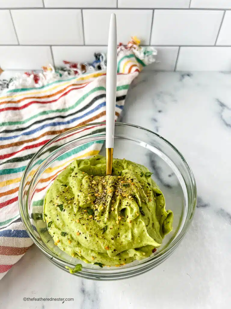 Serving spoon in a bowl of creamy avocado crema garnished with salt and cracked black pepper.