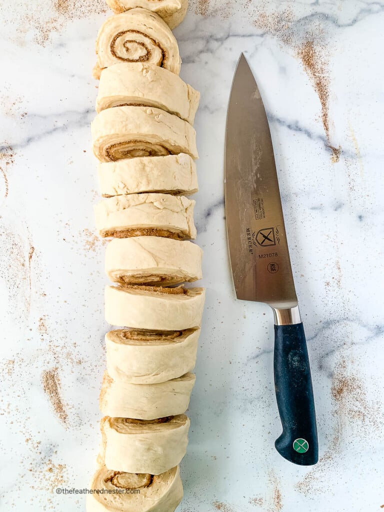Chef's knife next to slices of cinnamon roll dough on a marble countertop.