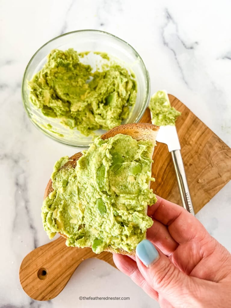 Bowl of green topping for toast in a small glass bowl and shmeared on a slice of bread.