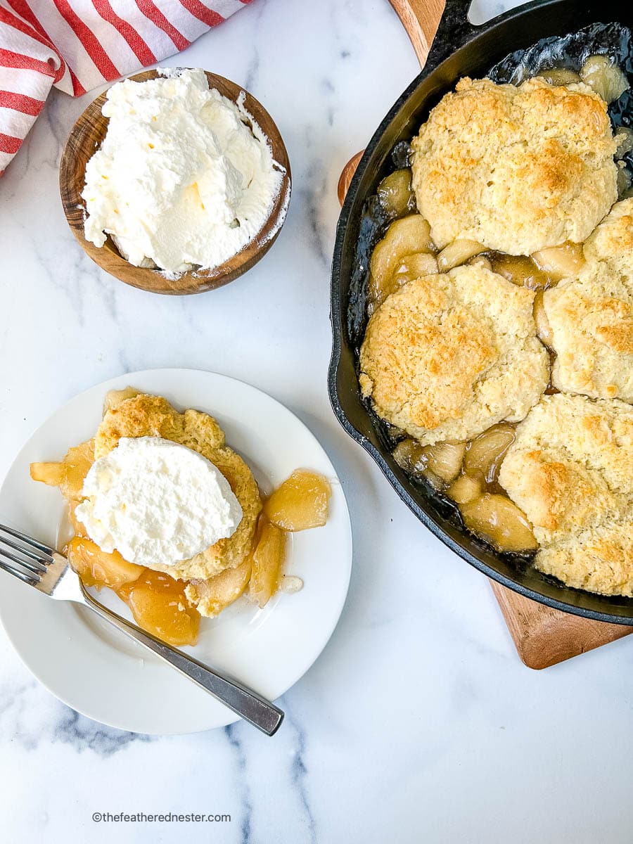 Serving of baked apple cobbler topped with whipped cream on a small dessert plate.
