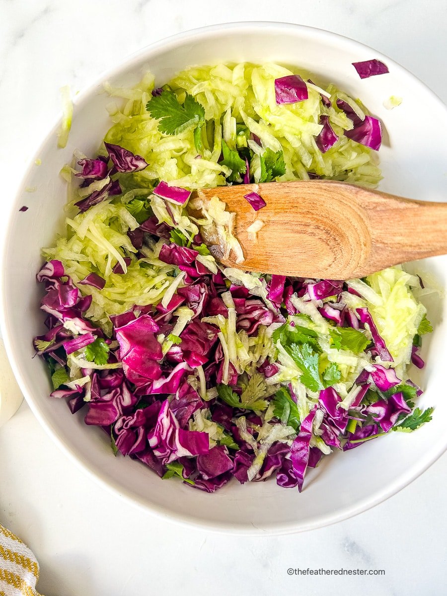 Using wooden spoon to combine shredded cucumbers and chopped red cabbage in a mixing bowl.