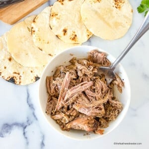 Shredded meat in a small white bowl next to charred tortillas.