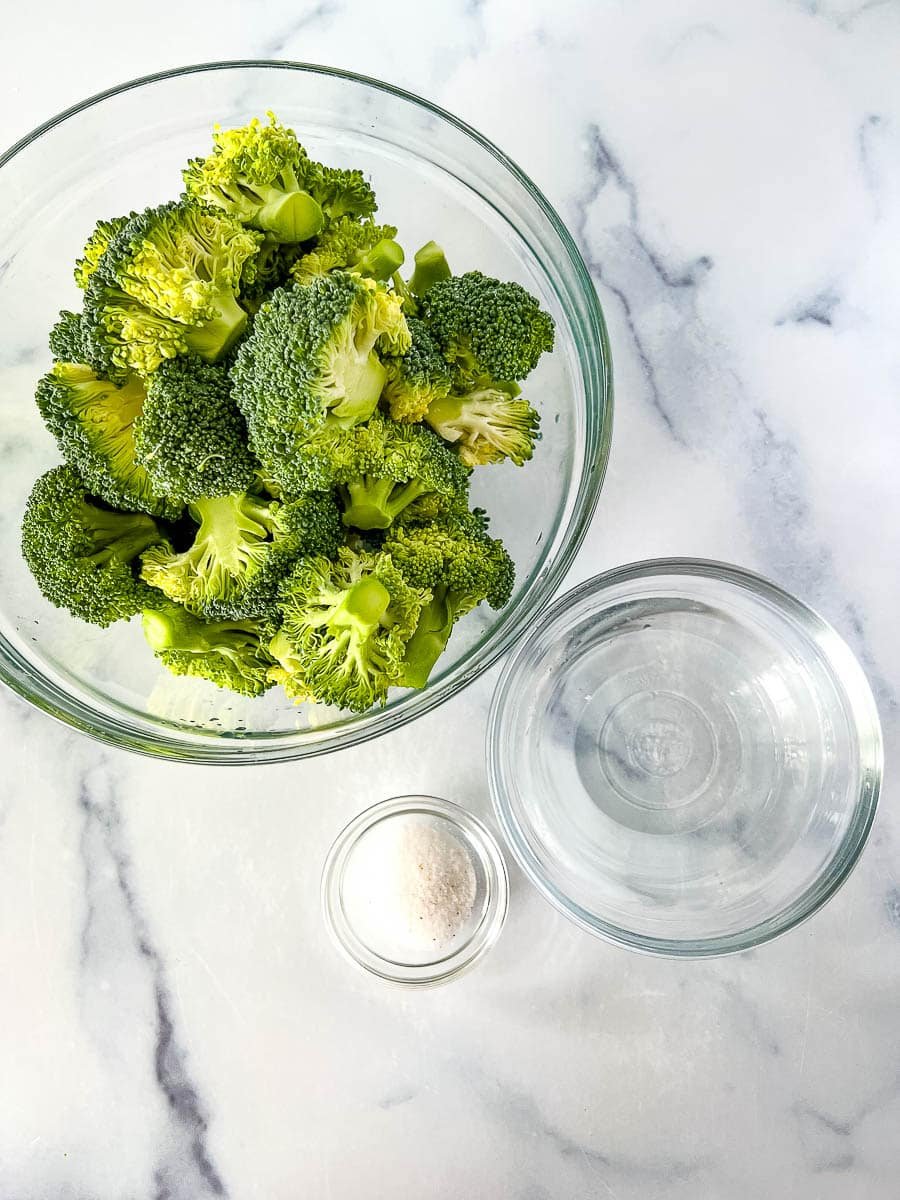 Raw broccoli next to a bowl with water and a smaller bowl of seasoning to make broccoli taste good.