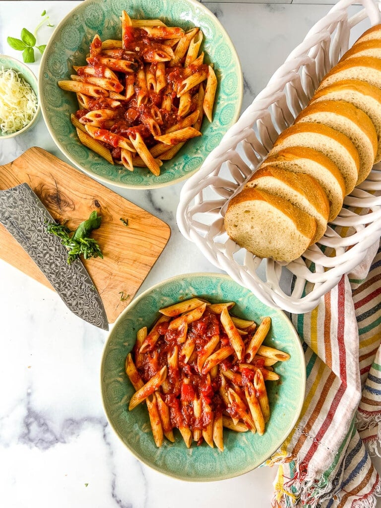 Two plated servings of penne pasta with meatless tomato basil pasta sauce next to a white basket with sliced French bread.