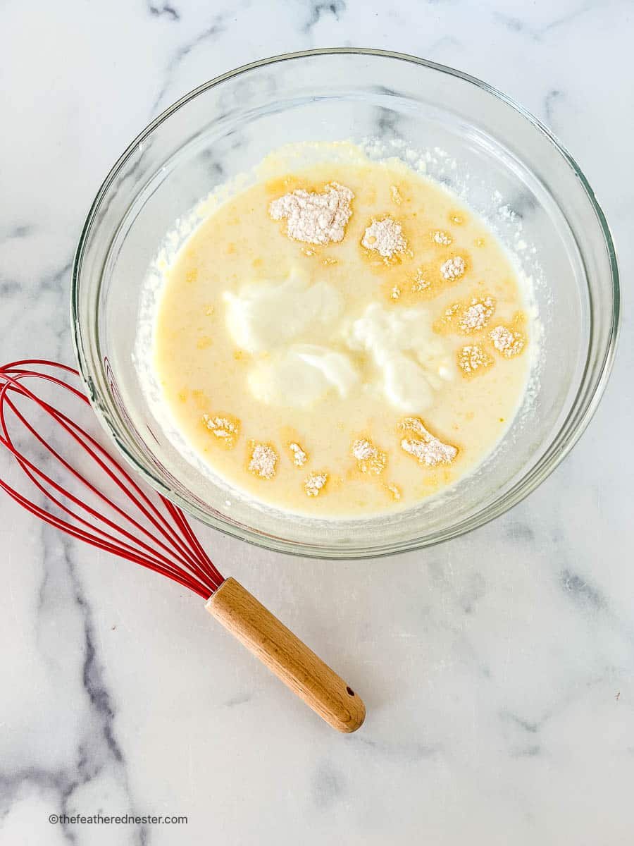 Red whisk sitting next to a clear mixing bowl filled with yogurt, dry pudding mix and milk.