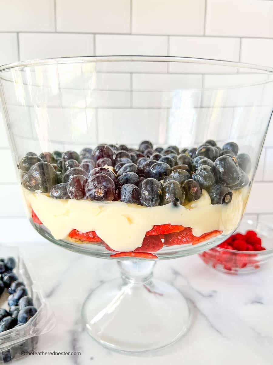 Assembling cheesecake salad with layers of fruit and pudding in a large glass bowl.