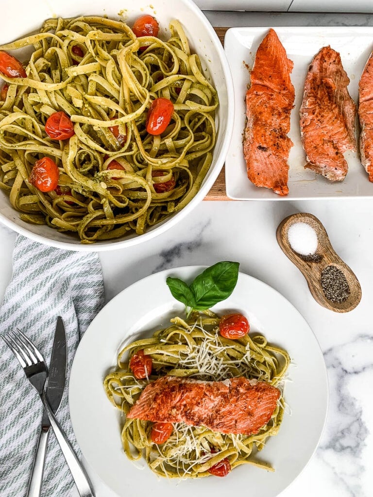 Salmon pesto pasta on a dinner plate. Bowl with pasta and a platter of baked fish are above the plate.