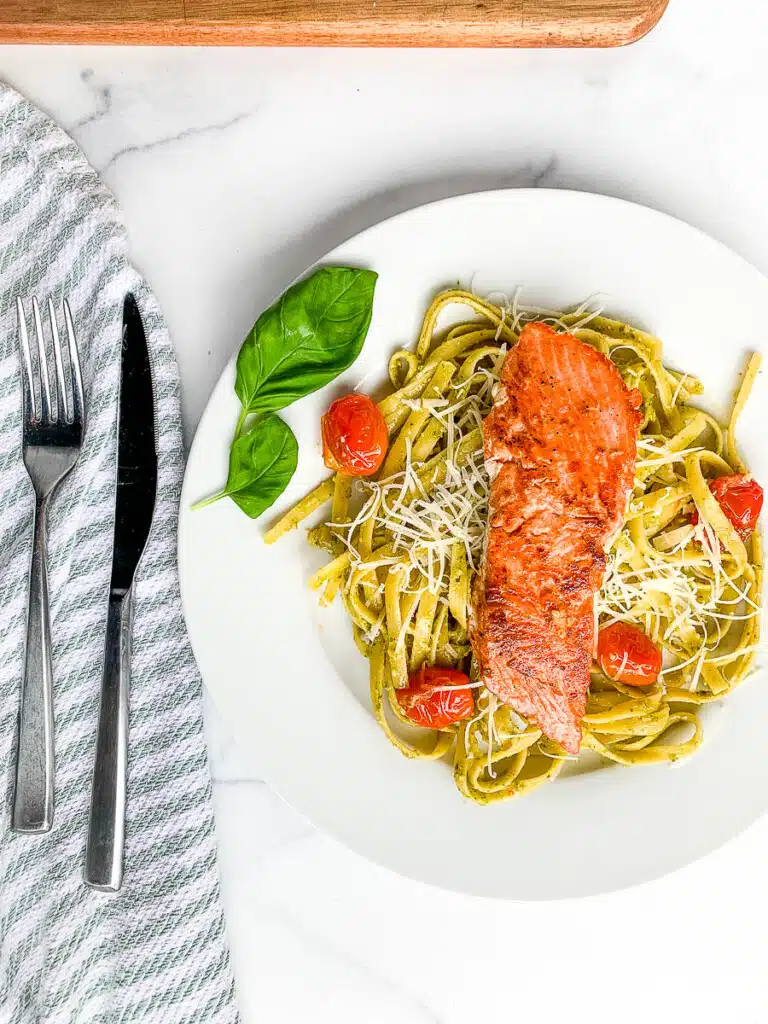 Fork and butter knife on a gray and white striped napkin next to salmon fettuccine on a white dinner plate.